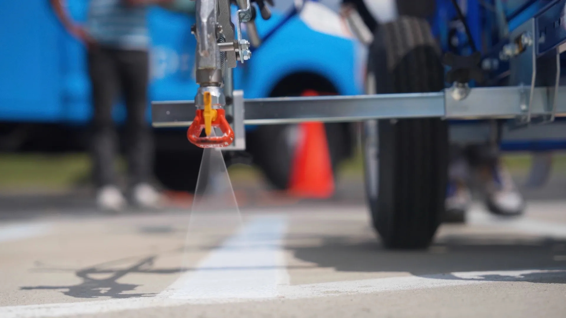 Appell striping line striping franchise excellence A close-up of a road striping machine spraying white paint on the pavement, with a blue truck and orange cone in the