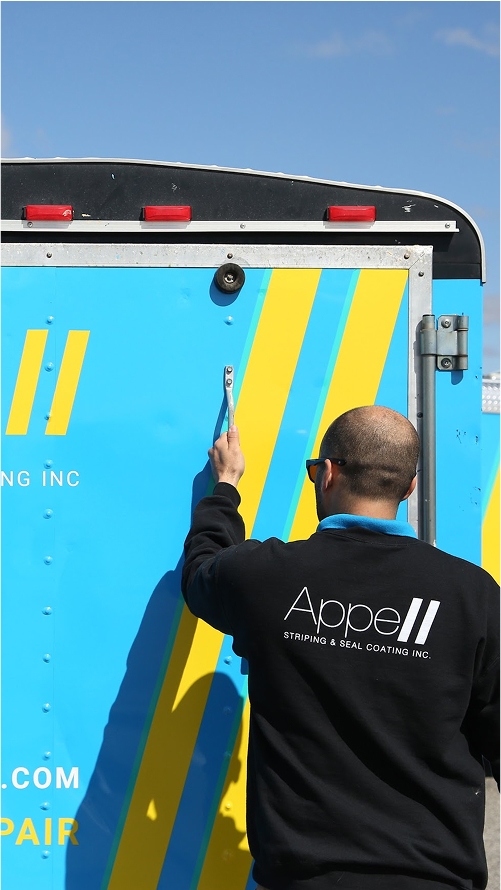 Why us Man in black jacket with 'AppeII Stripping & Seal Coating Inc.' logo opening the door of a blue and yellow truck.